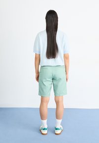 Top azul claro de manga corta con un acabado texturizado; pantalones cortos verde menta; calcetines blancos; zapatillas con un toque verde; de pie contra un fondo sencillo.