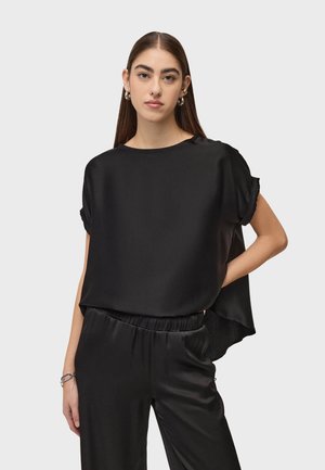 Kobieta ubrana w luźną, czarną satynową bluzkę z krótkim rękawem i pasujące spodnie stoi z jedną ręką na biodrze na jednolitym tle.