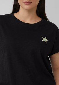 Zwarte short-sleeve t-shirt gemaakt van zachte stof, met een kleurrijke sterrenvormige geborduurde patch op de linkerkant van de borst.