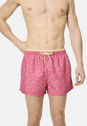 Torso masculino vestido con pantalones cortos de baño rosa con pequeños patrones de caballitos de mar azules y amarillos y un cordón amarillo, de pie contra un fondo liso.