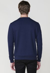 Hombre con un suéter de manga larga azul marino texturizado y pantalones negros, visto desde atrás contra un fondo claro.