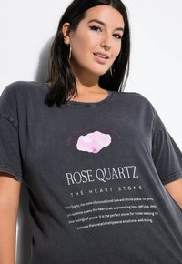 Donkergrijze oversized t-shirt met een roze afbeelding van een rozenkwarts steen en tekst die de eigenschappen beschrijft. Korte mouwen, ronde hals.
