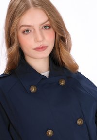 Marineblauwe double-breasted trenchcoat met grote gouden knopen, voorzien van een brede kraag en een gladde, lichte stof.