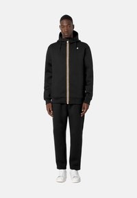 Sort zip-up hoodie med guld- og hvidstribe, der har en kangaroo-lomme og hætte med snøre. Sammen med matchende sorte bukser og hvide sneakers.