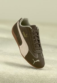 Ciemnobrązowe sneakersy Puma z białym paskiem, sznurowadłami i logo na teksturowanej jasnej powierzchni.