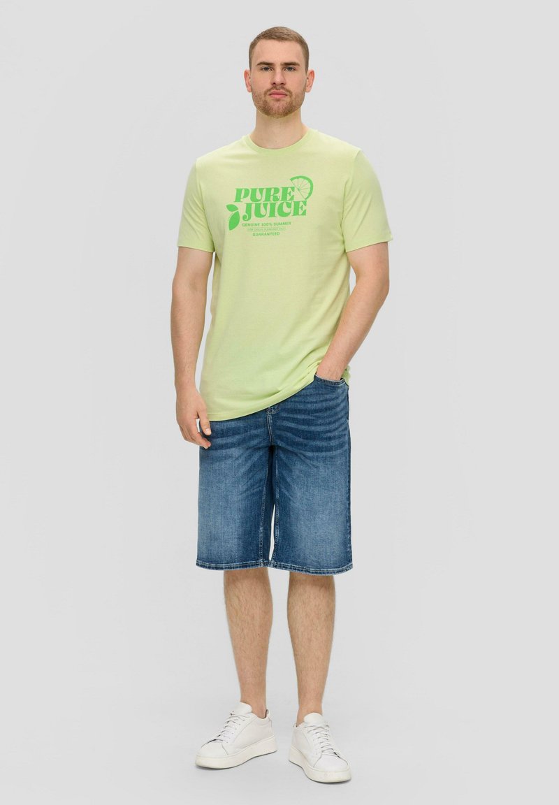 Lichtgroen T-shirt met "PURE JUICE" grafische opdruk in donkergroen. Gecombineerd met blauwe denim shorts en witte sneakers. Casual, ontspannen pasvorm.