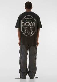 Svart oversized t-skjorte med grafisk design på baksiden, med et hodeskallemotiv og ordet "RODEO". Kombinert med mørke cargo-bukser.