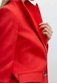 Una persona con capelli lunghi indossa un blazer rosso su misura, aggiustando il colletto con una mano su uno sfondo neutro.
