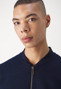 Sweat-shirt zippé navy avec col montant et tissu texturé. Le modèle présente une peau lisse et une expression neutre.
