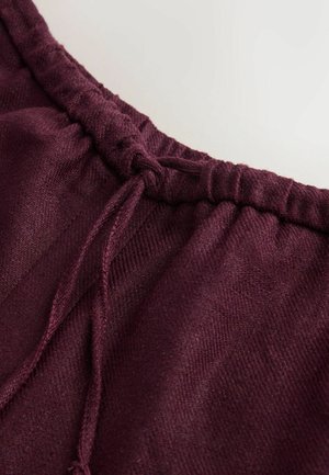 Nahaufnahme eines dunkelvioletten Stoffbunds mit elastischem Band und gebundenem Kordelzug vor einfarbigem Hintergrund.