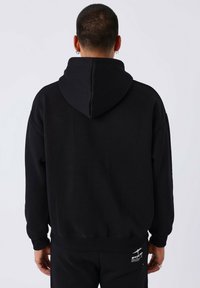 Man met kort haar en oorbellen, gekleed in een zwarte hoodie en zwarte broek, gezien vanaf achteren tegen een eenvoudige witte achtergrond.