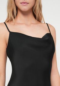 Μαύρο σατέν καμπάνα με λεπτές τιράντες, ελαφρύ V-ντεκολτέ και λεία υφή, αναδεικνύοντας έναν κομψό, μινιμαλιστικό σχεδιασμό.
