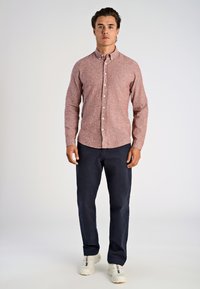 Man som står vänd mot kameran, iklädd en röd långärmad skjorta med knappar, mörkblå byxor och vita sneakers framför en enkel bakgrund.