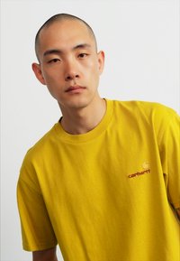 Geel katoenen T-shirt met een ronde halslijn, korte mouwen en een klein rood Carhartt-logo aan de linkerkant. Gladde textuur en casual pasvorm.
