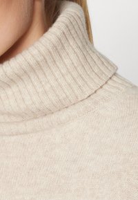 Gros plan d'une personne portant un pull à col roulé côtelé de couleur crème douce, avec un focus sur la zone du cou et des épaules.