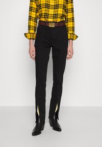 Persona con una camicia a quadri gialla, pantaloni neri con fessure frontali, cintura marrone e stivali neri con tacco, in piedi su uno sfondo bianco.