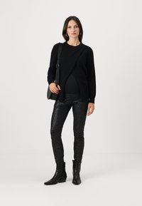 Svart tröja med lös passform, kombinerad med svarta texturerade leggings och ankelstövlar. Modellen bär en svart väska och står mot en vit bakgrund.