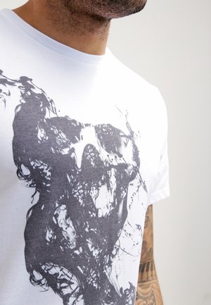 Mand iført hvid T-shirt med abstrakt sort kraniedesign, delvist synlig tatoveret arm, neutralt baggrund.