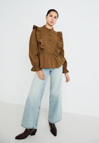 Blusa a quadri marrone con dettagli a ruches, chiusura a bottoni e maniche lunghe, abbinata a jeans a gamba larga blu chiaro e stivali con tacco marroni.
