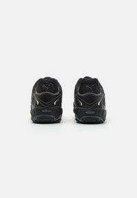 Des chaussures de sport noires avec une tige texturée, un col rembourré et une semelle épaisse avec un logo proéminent au niveau du talon. Accents réfléchissants présents.