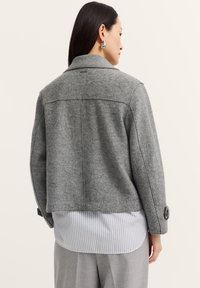 Grau melierte Wolljacke mit einem Kragen, Rückennähten und Knopfmanschetten, kombiniert mit einem hell gestreiften Hemd darunter.