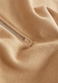 Beige gebreide stof met een gladde textuur, voorzien van een zilveren ritsdetail. De stof toont zachte plooien en subtiele schaduwen.