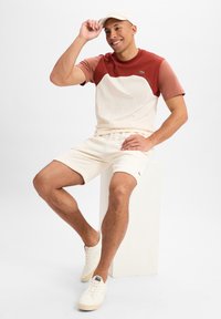 Beige en roestkleurig shirt met korte mouwen, crème korte broek en bijpassende witte sneakers. Glad textiel met een klein logo op het shirt en de korte broek.