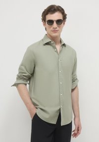 Uomo con occhiali da sole tondi neri, camicia verde salvia con bottoni e maniche rimboccate, e pantaloni neri, in piedi contro uno sfondo bianco.
