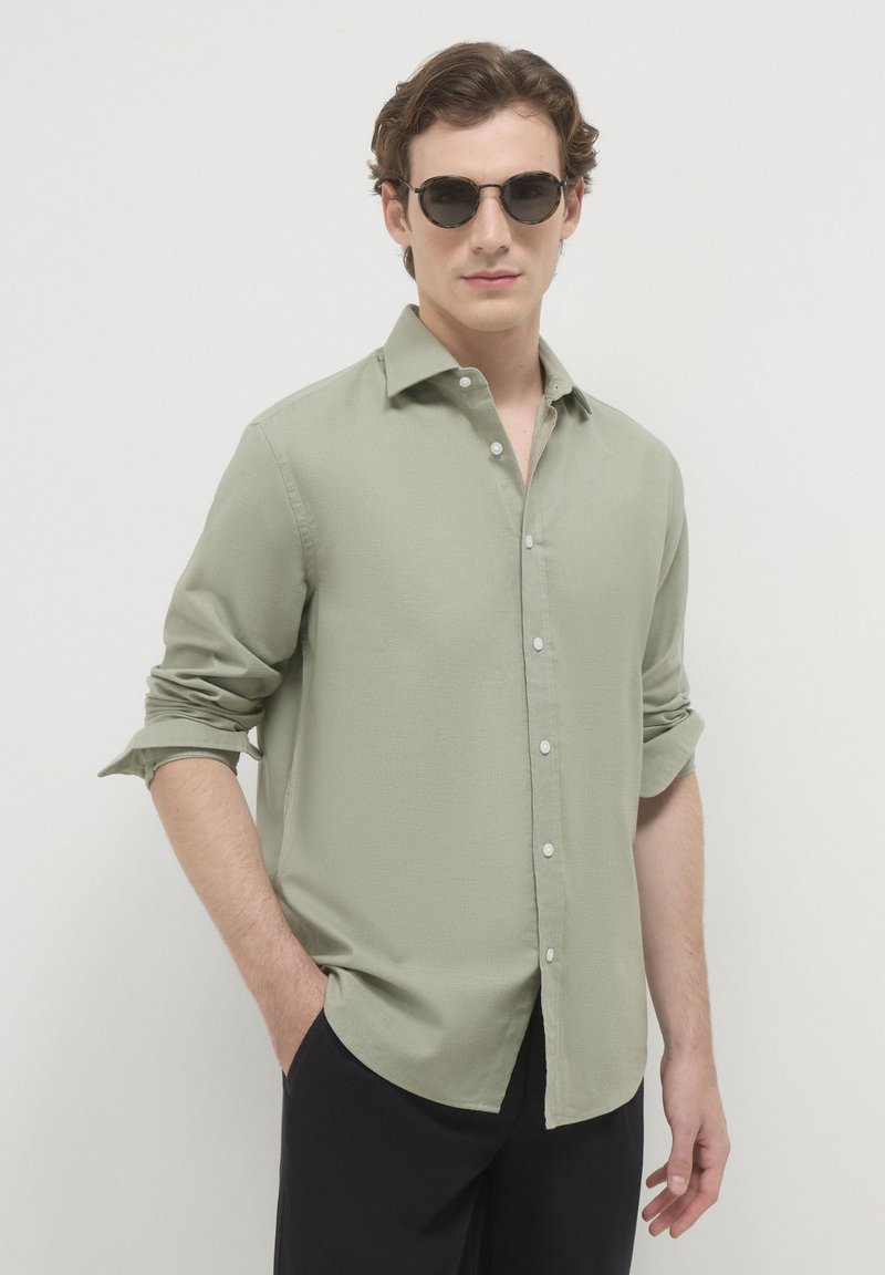 Uomo con occhiali da sole tondi neri, camicia verde salvia con bottoni e maniche rimboccate, e pantaloni neri, in piedi contro uno sfondo bianco.