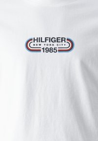 Bílá látka s textem "HILFIGER NEW YORK CITY 1985" v černé barvě, doplněná zakřivenými červenými a modrými pruhovanými liniemi po každé straně.