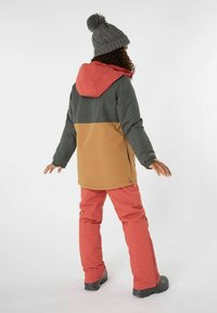 Giacca da esterno con un design a blocchi di colore verde e sabbia, cappuccio rosso, abbinata a pantaloni da neve rosa. Presenta polsini elasticizzati e dettagli con zip.