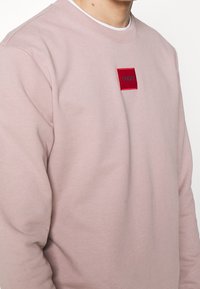 Sudadera rosa clara hecha de una tela suave, con un parche cuadrado rojo que tiene el texto "HUGO" centrado en el pecho. Diseño de cuello redondo acanalado.