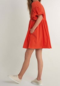 Vestido corto naranja brillante con cuerpo acanalado y mangas abullonadas, que cuenta con dos bolsillos laterales. Combinado con zapatillas beige.