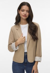 Beige blazer met een getailleerde pasvorm, met opgerolde mouwen met witte strepen, gecombineerd met een witte T-shirt en blauwe spijkerbroek. Soepele textuur.