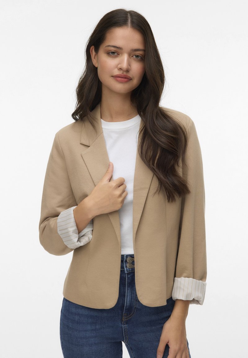 Beige blazer met een getailleerde pasvorm, met opgerolde mouwen met witte strepen, gecombineerd met een witte T-shirt en blauwe spijkerbroek. Soepele textuur.