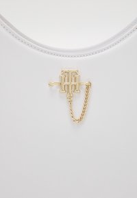 Biała torba o gładkiej skórzanej fakturze, z złotym akcentem logo oraz łańcuszkiem jako detal ozdobny.