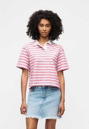 Kaaslastega naine, kes kannab roosa, valge ja sinise triibulise polo särgi ning kulunud helerohelist teksaseelikut valge tausta ees.