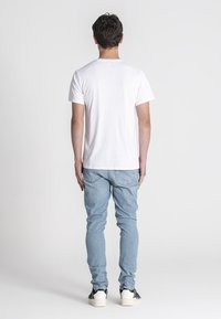 Camiseta blanca de manga corta vista desde la espalda, combinada con jeans de denim azul claro y zapatillas blancas con acentos negros. Textura de tela suave.