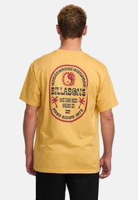Gele katoenen t-shirt met een cirkelvormige rode en zwarte afbeelding op de achterkant met tropische motieven, merktekst en een zonontwerp.