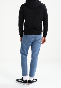 Persona che indossa un hoodie nero, jeans azzurri chiari, calze bianche e sneakers nere, in piedi con la schiena rivolta verso la fotocamera su uno sfondo bianco.