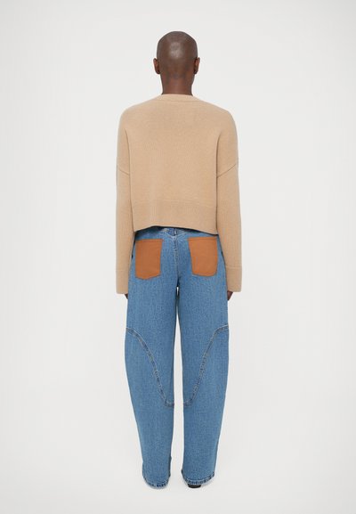 Pulover scurt de culoare maro deschis cu tiv ondulat, asociat cu blugi largi albaștri, având petice din piele bej pe buzunarele din spate și cusături curbate.