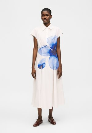 Γυναίκα που φοράει ένα λευκό μίντι φόρεμα με κοντά μανίκια και μεγάλο μπλε λουλουδάτο σχέδιο στο μπροστινό μέρος, συνδυασμένο με σκούρα καφέ παπούτσια.