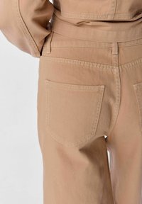Beige bomullbyxor med slät textur, med två bakfickor och synliga sömnadsdetaljer längs midjan och sömmarna.