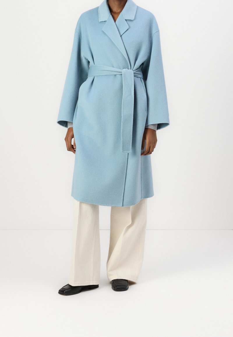 Personne portant un manteau en laine bleu clair ceinturé sur un pantalon crème à jambes larges et des chaussures noires à bout carré sur fond blanc.