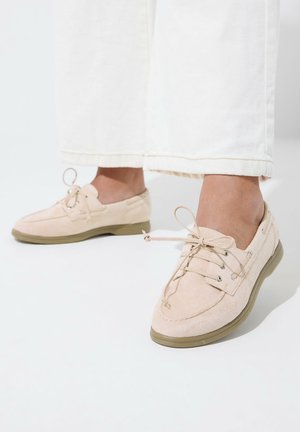 Jalat, joissa on vaaleanbeiget mokkakengät, joissa on kumipohjat ja nauhat, yhdistettynä valkoisiin leveälahkeisiin housuihin.