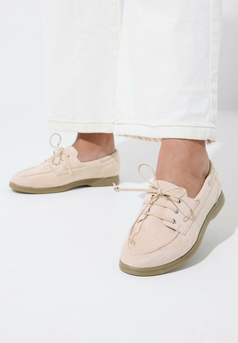 Pieds chaussés de chaussures à lacets en daim beige clair avec semelles en caoutchouc, assorties à un pantalon blanc à jambes larges.