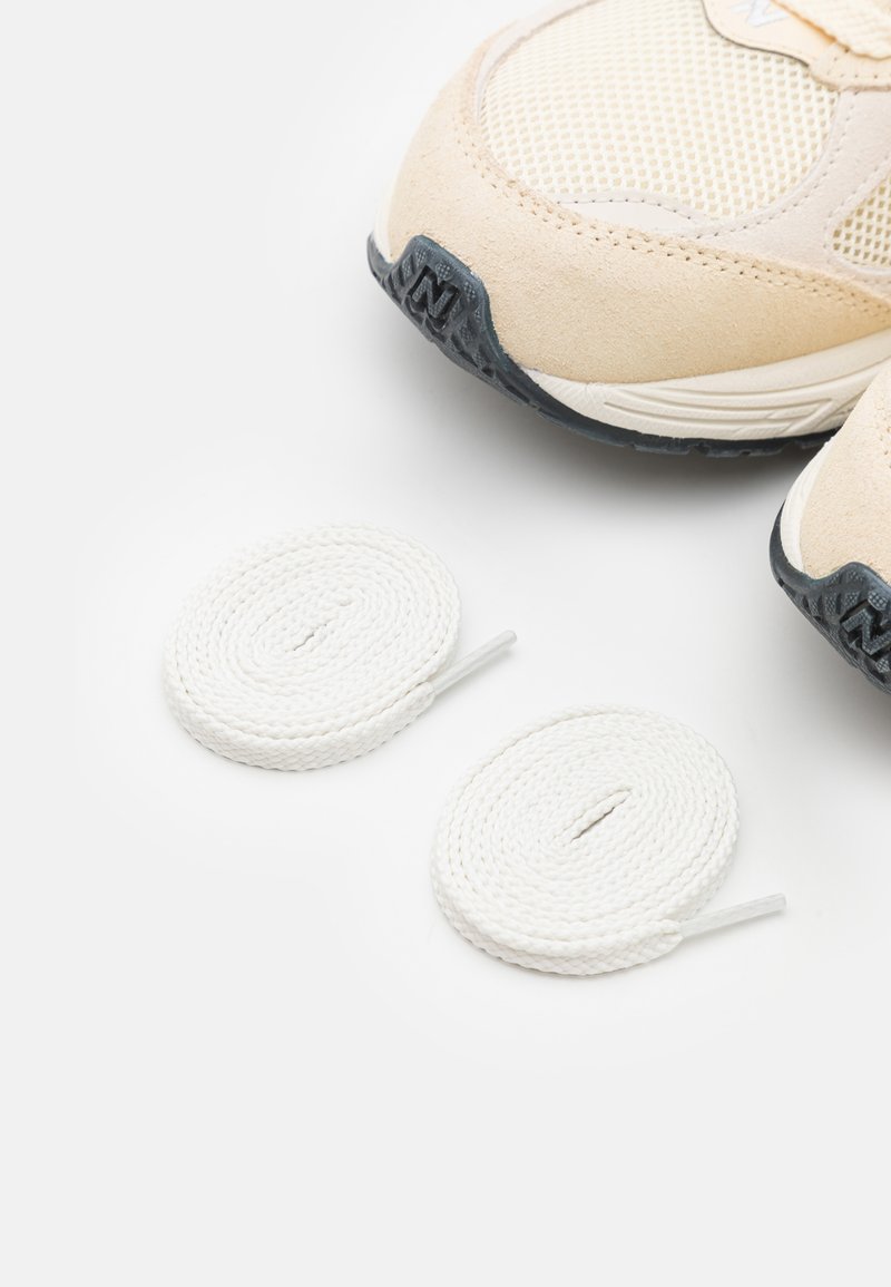 Scarpe sportive beige con tomaia in rete e dettagli in suede, abbinate a due lacci bianchi, esposte su uno sfondo neutro.