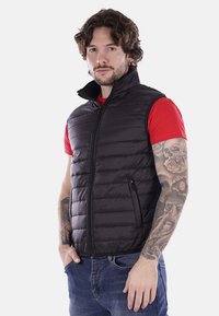 Gilet in piuma nero con collo alto, chiusura con zip e tasche laterali; presenta una texture trapuntata e una vestibilità aderente, indossato sopra una maglietta rossa.