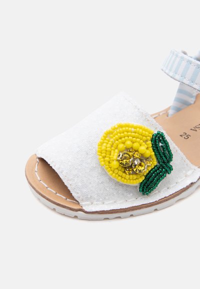 Παιδικό σανδάλι σε λευκό χρώμα, με ανοιχτή μύτη, λουράκι στον αστράγαλο και κίτρινο διακοσμητικό λουλούδι από χάντρες με πράσινα φύλλα στο μπροστινό λουράκι.