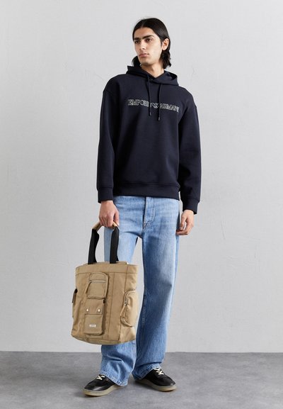 Borsa a spalla in canvas beige con manici neri, tasche frontali e logo. Indossata con una felpa blu navy e jeans azzurri, in piedi contro uno sfondo grigio.
